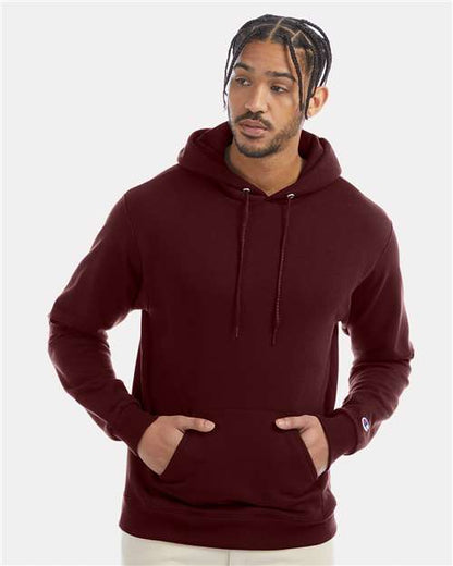 A man models the maroon Champion S700 hoodie, crafted from recycled polyester, standing against a plain light background with his hands in the pockets and gazing slightly to the side.
