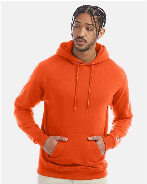 A man with braided hair wears a Champion S700 orange hoodie, made from recycled polyester, standing against a plain light gray background with his hands in the pocket, looking slightly to the side.