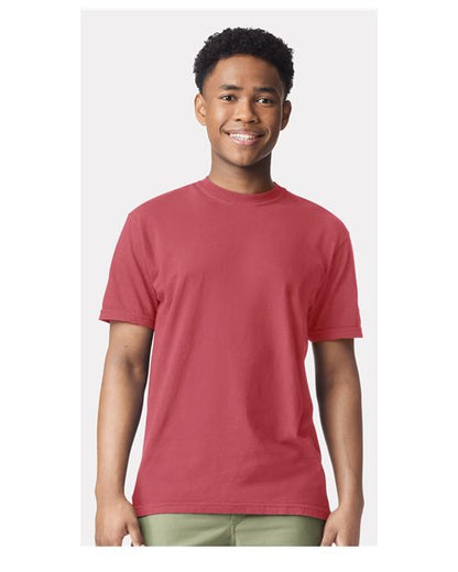 A young person with short curly hair smiles while wearing the Comfort Colors 1717 garment-dyed t-shirt in red, made from sustainable cotton, and light green pants, standing against a light gray background.