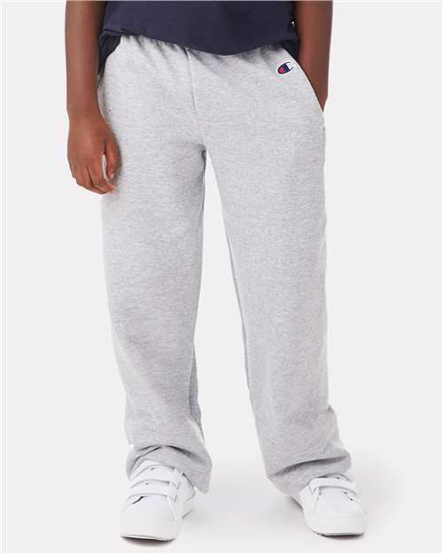 A person in light gray Champion P890 cotton-polyester sweatpants with side seam pockets, a dark shirt, and white sneakers stands with hands at their sides. The Champion logo is visible on the left hip of the pants.