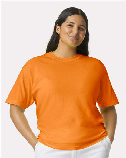 A person with long dark hair wears the Comfort Colors 1717 garment-dyed orange short-sleeve t-shirt (OEKO-TEX certified) and white pants, smiling with hands in pockets against a plain light background.
