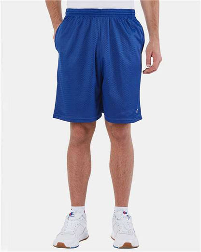 A person wearing Champion S162 athletic shorts stands with hands in pockets against a plain white background, showing only the lower torso and legs. The shorts are made of polyester mesh and paired with white sneakers.