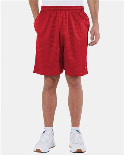 A person wears Champion S162 athletic shorts with pockets and an elastic waistband, paired with white sneakers and a white shirt. The red polyester mesh shorts stand out against a plain light background as the person stands hands in pockets.
