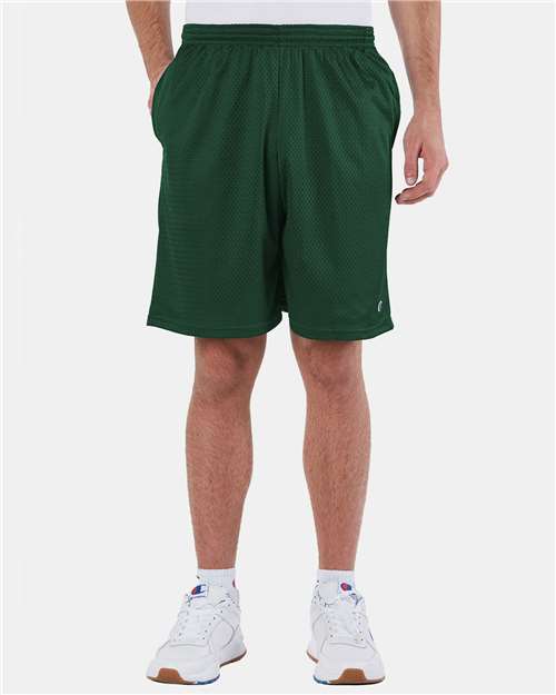 A person wearing Champion S162 dark green polyester mesh athletic shorts, a white shirt, and white athletic shoes stands against a plain light background with hands in their pockets. The persons head is not visible.