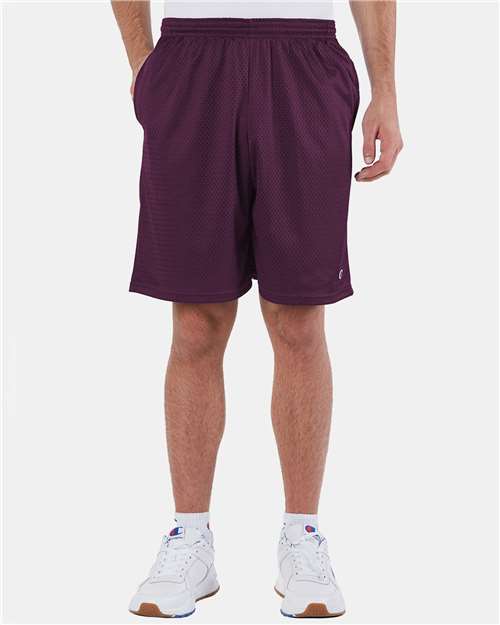 A person stands against a plain light background wearing maroon Champion S162 athletic shorts made from polyester mesh, a white t-shirt, and white sneakers. Their face is not visible and their hands are relaxed at their sides.