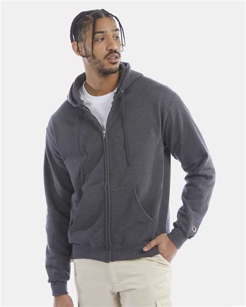A man in a dark gray Champion S800 hoodie made from recycled polyester stands with one hand in his pocket over a white tee and light pants, looking to the side against a plain light background.