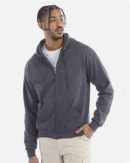 A man in a dark gray Champion S800 hoodie made from recycled polyester stands with one hand in his pocket over a white tee and light pants, looking to the side against a plain light background.