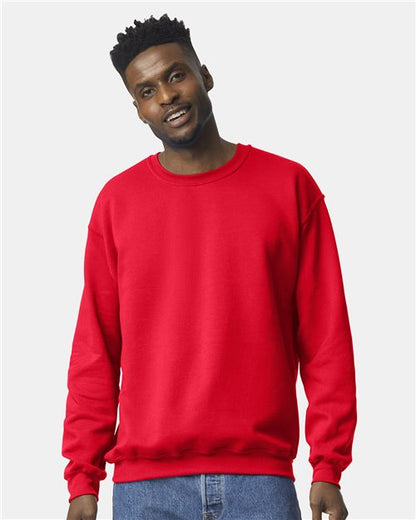 A man with short, curly hair wears a bright red Gildan 18000 sweatshirt and blue jeans, standing against a plain light gray background and smiling slightly.