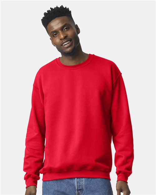 A man with short, curly hair wears a bright red Gildan 18000 sweatshirt and blue jeans, standing against a plain light gray background and smiling slightly.