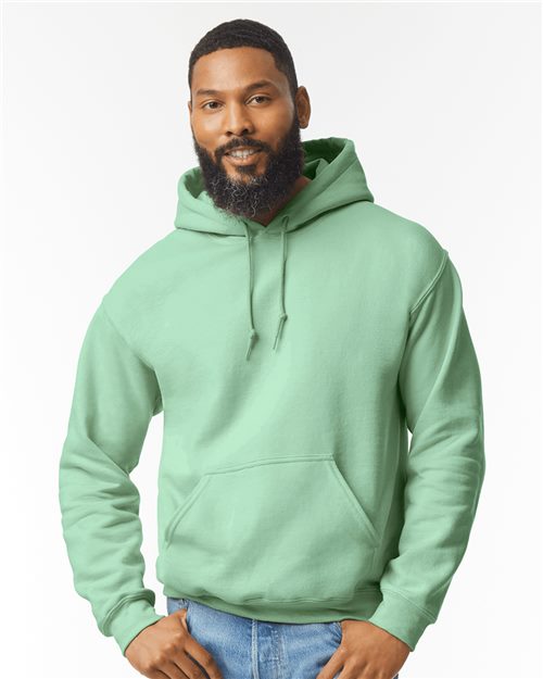 A man with a short beard and mustache wears a light green Gildan 18500 Heavy Blend hoodie and blue jeans, standing with his hands near his pockets against a plain white background.