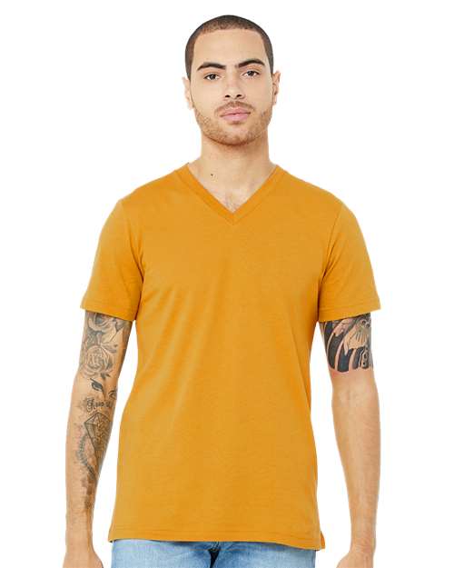 A person with short hair and tattooed arms wears the BELLA + CANVAS 3005 unisex V-neck in mustard yellow (Airlume combed cotton) and light blue jeans, standing against a plain white background.