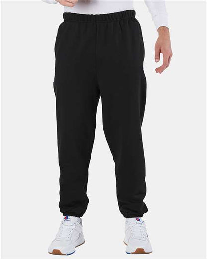 A person stands against a plain, light background wearing Champion RW10 black cotton-polyester jogger sweatpants with elastic cuffs, a white long-sleeve shirt, and white athletic shoes. The focus is on the lower half of the body.