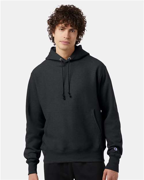 A young person with curly hair wears the black Champion S101 sweatshirt, featuring front pockets and drawstrings, standing against a plain light gray background.