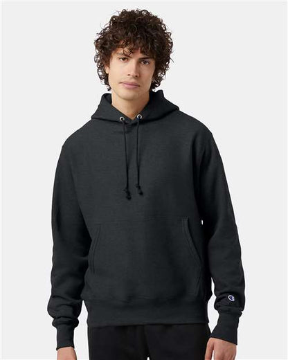 A young person with curly hair wears the black Champion S101 sweatshirt, featuring front pockets and drawstrings, standing against a plain light gray background.