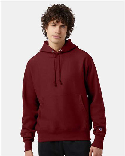 A young person with curly hair wears the Champion S101 sweatshirt, featuring a front pocket and drawstrings, standing against a plain light gray background.