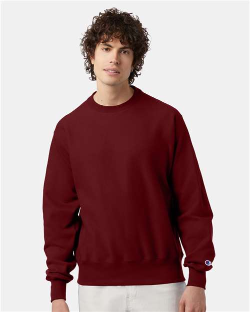 A young man with curly brown hair wears a maroon Champion S149 sweatshirt made from a cotton-polyester blend and light-colored pants, standing against a plain light gray background.