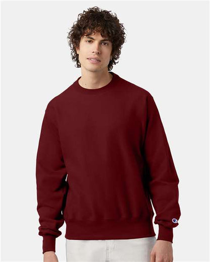 A young man with curly brown hair wears a maroon Champion S149 sweatshirt made from a cotton-polyester blend and light-colored pants, standing against a plain light gray background.