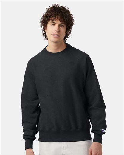 A young man with curly hair wears a plain black Champion S149 sweatshirt and light-colored pants, standing against a light gray background.