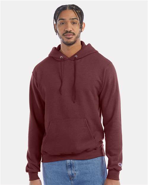 A man stands facing forward against a plain light gray background, wearing a maroon Champion S700 hoodie with drawstrings and a front pocket, made from recycled polyester, paired with light blue jeans.