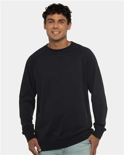 A young man with short, curly dark hair smiles in a Next Level 9000 plain black cotton polyester fleece crewneck sweatshirt with a classic v-notch neck, paired with light blue jeans against a light gray background.