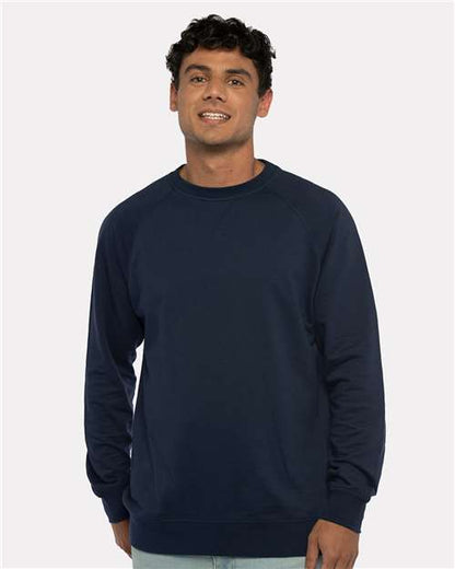A man with short curly dark hair wears the Next Level 9000 navy blue crewneck sweatshirt with a classic v-notch neck and light blue jeans, standing and smiling against a plain light background.