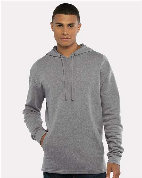 A young man stands facing forward, one hand in his pocket, smiling softly while wearing the Next Level 9300 light gray cotton-polyester fleece hoodie with drawstrings against a plain, light background.