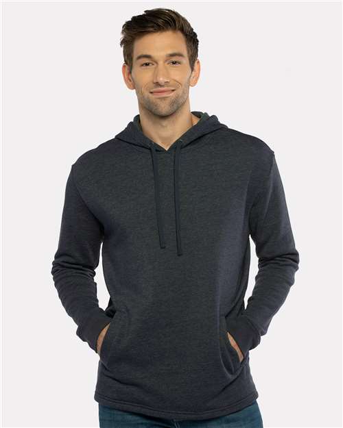 A man wears the Next Level 9300 hoodie, made from cotton polyester fleece, standing against a light background with hands in the front pocket, facing the camera and smiling softly.