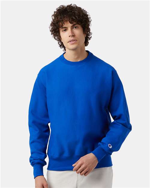 A person with curly brown hair wears a bright blue Champion S149 sweatshirt and light-colored pants, standing against a plain light background.