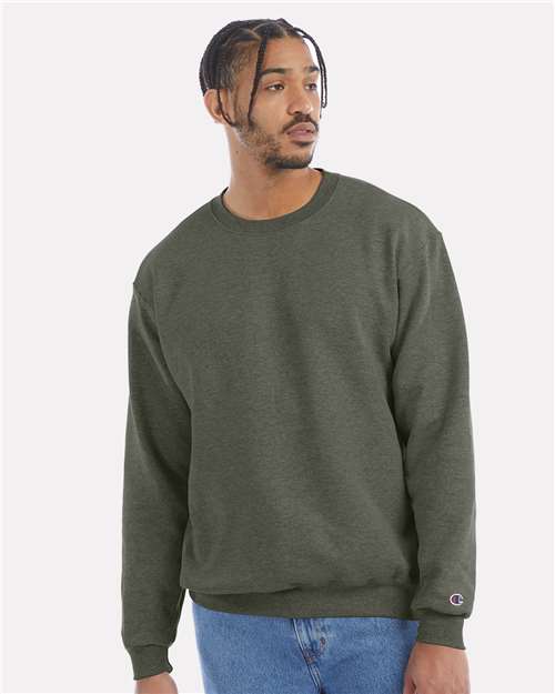 A man with short braids and a trimmed beard wears a Champion S600 olive green crewneck sweatshirt and blue jeans, looking to his left against a plain light background.