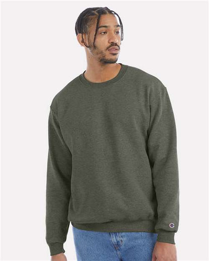 A man with short braids and a trimmed beard wears a Champion S600 olive green crewneck sweatshirt and blue jeans, looking to his left against a plain light background.