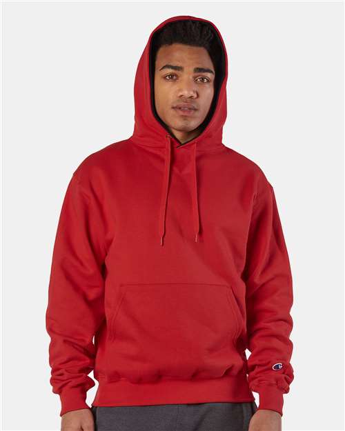 A person wearing a bright red Champion S171 cotton-polyester hoodie with the hood up stands against a plain light background. The sweatshirt has a pouch pocket, drawstrings, and an embroidered logo. They look directly at the camera.