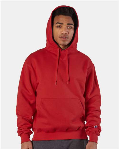 A person wearing a bright red Champion S171 cotton-polyester hoodie with the hood up stands against a plain light background. The sweatshirt has a pouch pocket, drawstrings, and an embroidered logo. They look directly at the camera.
