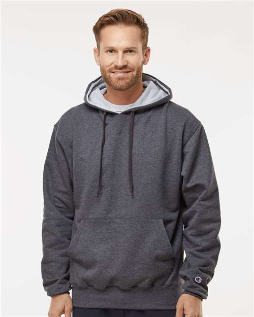 A man with short light brown hair and a beard is wearing the Champion S171 dark gray hoodie, featuring an embroidered logo and pouch pocket, standing against a plain light background and smiling slightly at the camera.