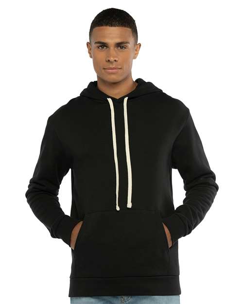 A young man in a Next Level 9303 black cotton-polyester hoodie with white drawstrings and a jersey-lined hood stands casually with hands in the front pocket, wearing blue jeans and looking straight ahead.