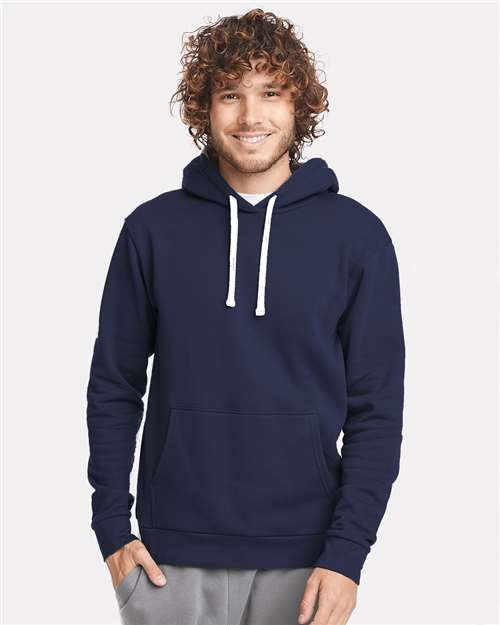 A smiling person with curly hair wears the Next Level 9303 navy blue cotton-polyester hoodie with white drawstrings, a jersey-lined hood, and front pocket, standing against a plain light background.