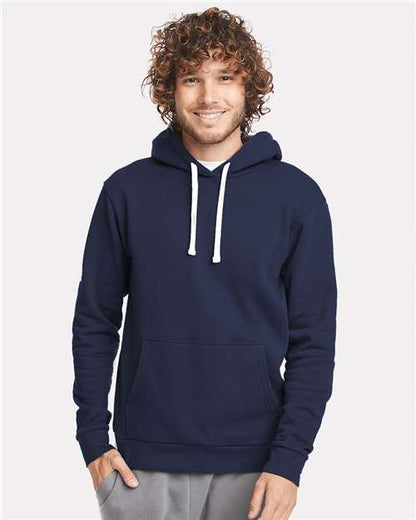 A smiling person with curly hair wears the Next Level 9303 navy blue cotton-polyester hoodie with white drawstrings, a jersey-lined hood, and front pocket, standing against a plain light background.