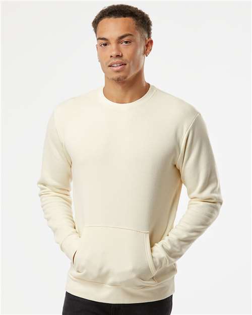 A young man with short curly hair wears the Next Level 9001 cream-colored, long-sleeve crewneck sweatshirt with front pockets, made from cotton/polyester fleece, standing against a plain white background.