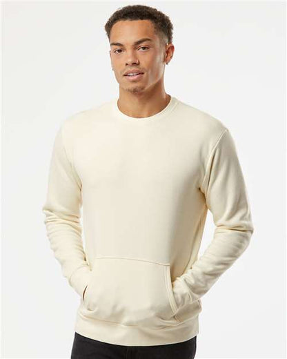 A young man with short curly hair wears the Next Level 9001 cream-colored, long-sleeve crewneck sweatshirt with front pockets, made from cotton/polyester fleece, standing against a plain white background.