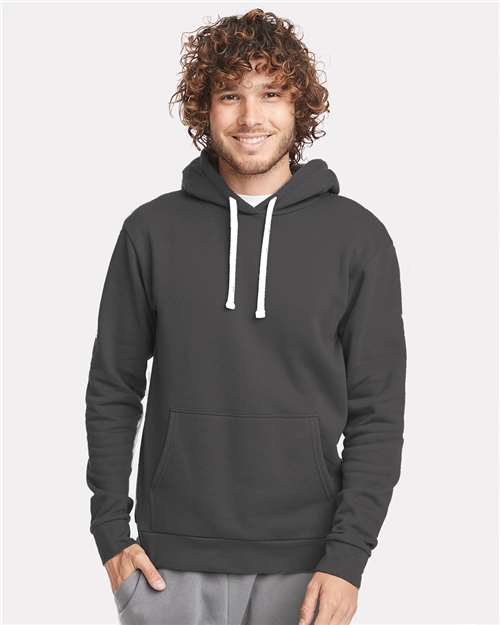 A smiling person with curly hair wears the Next Level 9303 dark gray cotton-poly hoodie with white drawstrings, front pocket, and soft jersey-lined hood, standing against a light background.