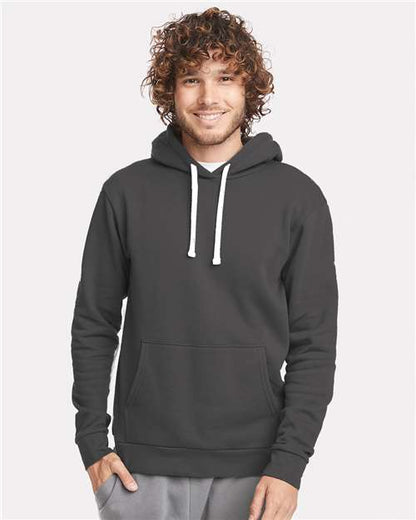 A smiling person with curly hair wears the Next Level 9303 dark gray cotton-poly hoodie with white drawstrings, front pocket, and soft jersey-lined hood, standing against a light background.