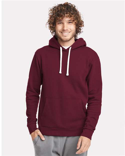 A person with curly hair is smiling and wearing the Next Level 9303 maroon cotton-polyester hoodie with jersey-lined hood, white drawstrings, and front pocket, paired with light gray pants against a plain light background.