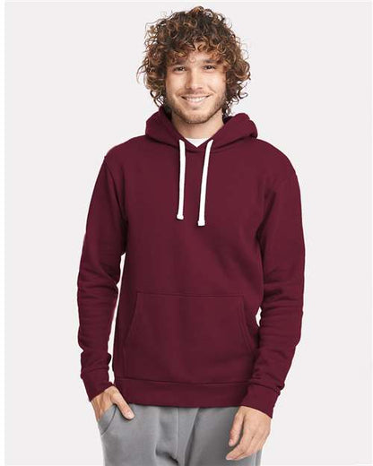 A person with curly hair is smiling and wearing the Next Level 9303 maroon cotton-polyester hoodie with jersey-lined hood, white drawstrings, and front pocket, paired with light gray pants against a plain light background.