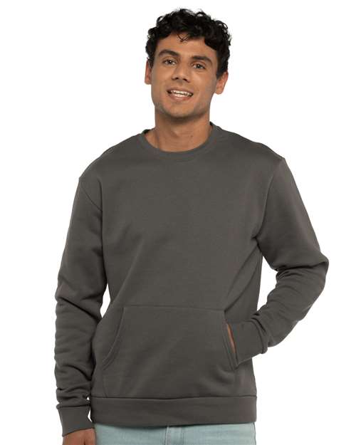 A young man with short curly hair smiles while wearing the Next Level 9001 dark gray unisex crewneck sweatshirt with a front pocket, one hand in the pocket, standing against a plain white background.