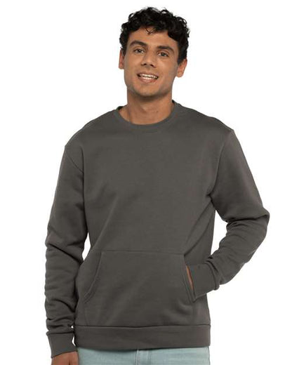 A young man with short curly hair smiles while wearing the Next Level 9001 dark gray unisex crewneck sweatshirt with a front pocket, one hand in the pocket, standing against a plain white background.