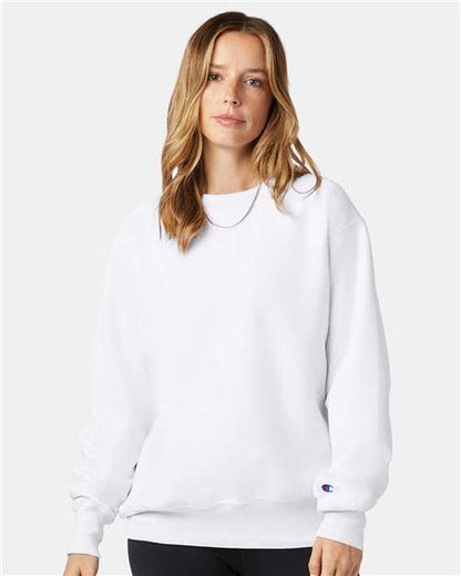 A woman with light brown hair wears a plain white Champion S149 sweatshirt and black pants, standing against a light background. The small sleeve logo highlights its cotton-polyester blend and sustainable manufacturing.