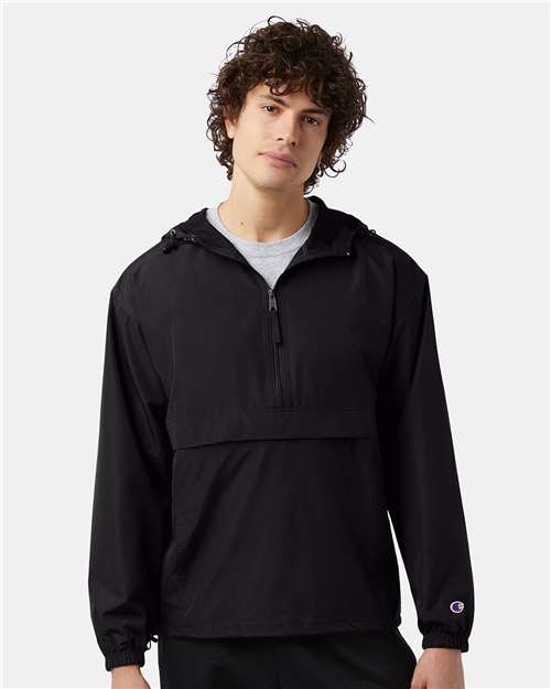 A young man with curly brown hair wears a Champion CO200 black, long-sleeve, hooded wind- and water-resistant jacket with a front half-zip and pocket, standing against a plain light gray background.