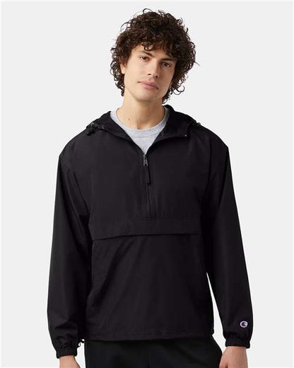 A young man with curly brown hair wears a Champion CO200 black, long-sleeve, hooded wind- and water-resistant jacket with a front half-zip and pocket, standing against a plain light gray background.