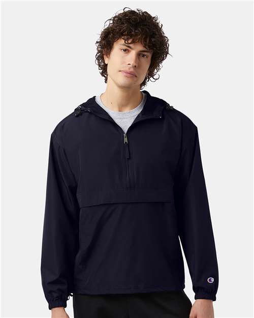 Curly-haired young man in a black Champion CO200 jacket, featuring wind- and water-resistance, front zipper, and large pocket, stands against a plain light background.