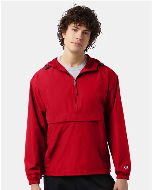 A young man with curly brown hair wears the Champion CO200 bright red wind- and water-resistant jacket with a front pocket, half-zip, and hood, standing against a plain light background.