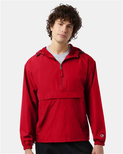 A young man with curly brown hair wears the Champion CO200 bright red wind- and water-resistant jacket with a front pocket, half-zip, and hood, standing against a plain light background.